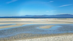 Eine Küstenlandschaft mit blauem Himmel, fernen Bergen und einem Strand mit flachem Wasser und Kieselsteinen.