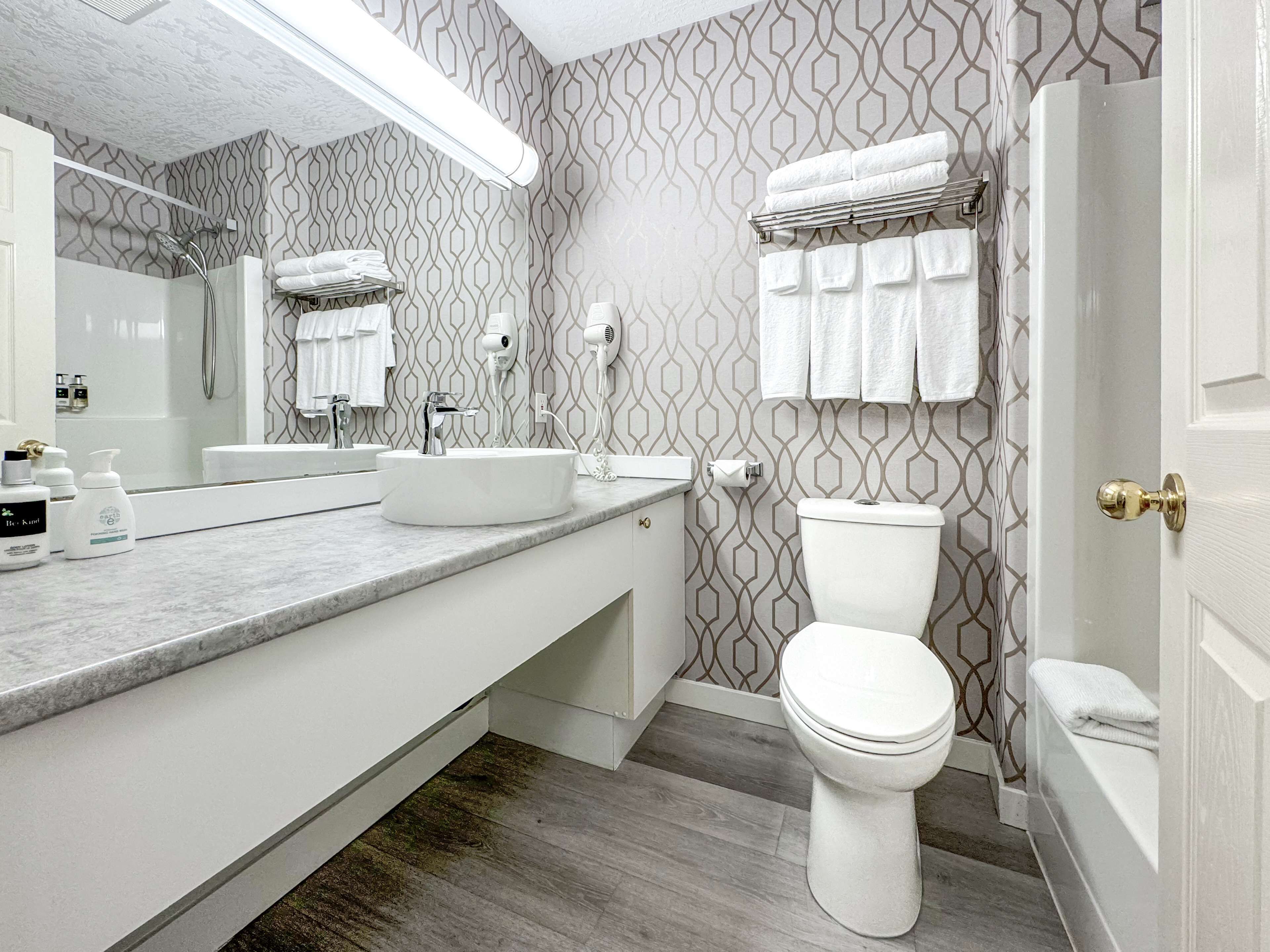 Un baño moderno con un lavabo, tocador y retrete blancos. Hay un perchero sobre el retrete y un cabezal de ducha en la pared.