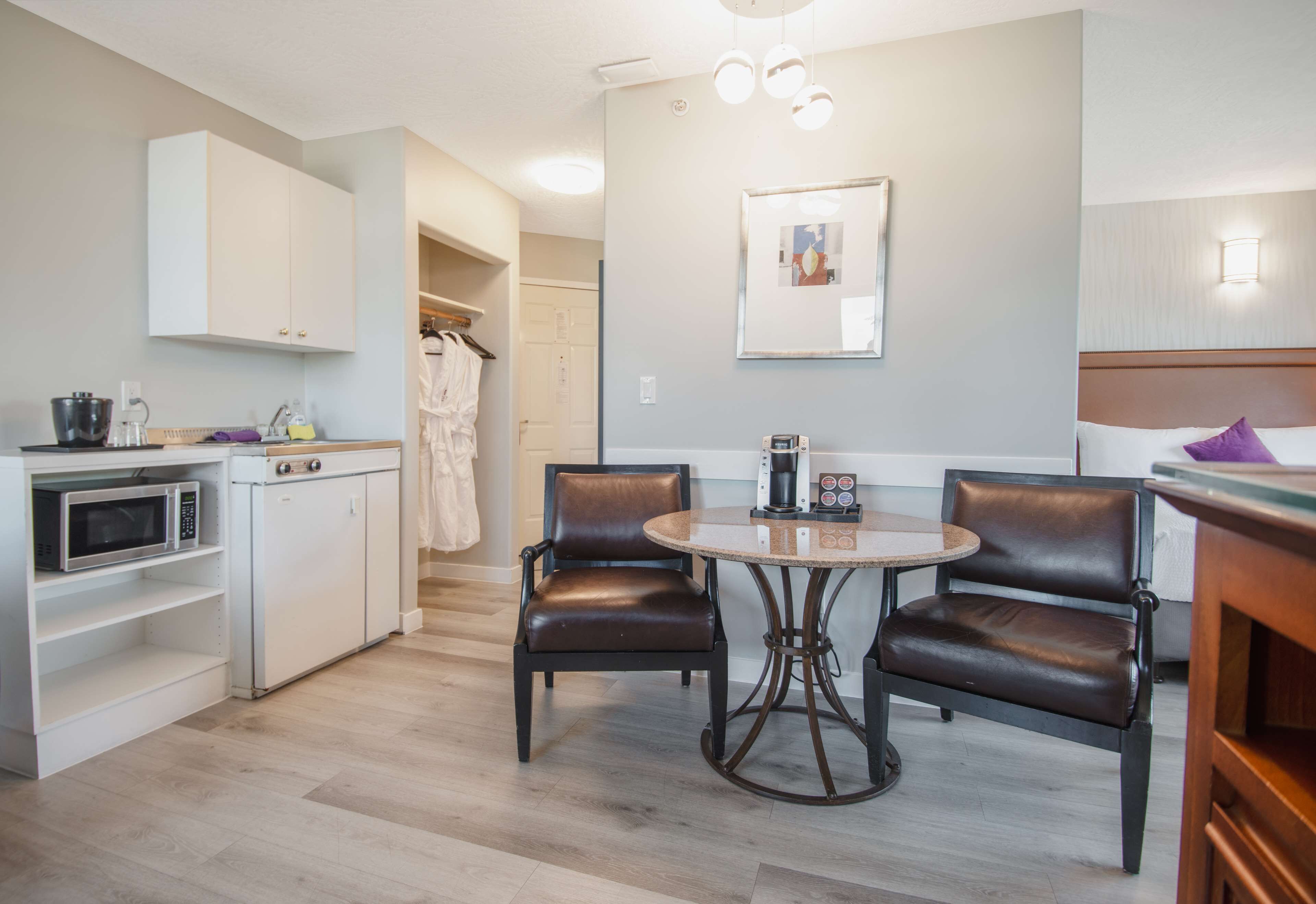 A compact kitchen with a microwave, sink, and cabinets. There is a dining area with two chairs and a table, and a coat rack in the background.