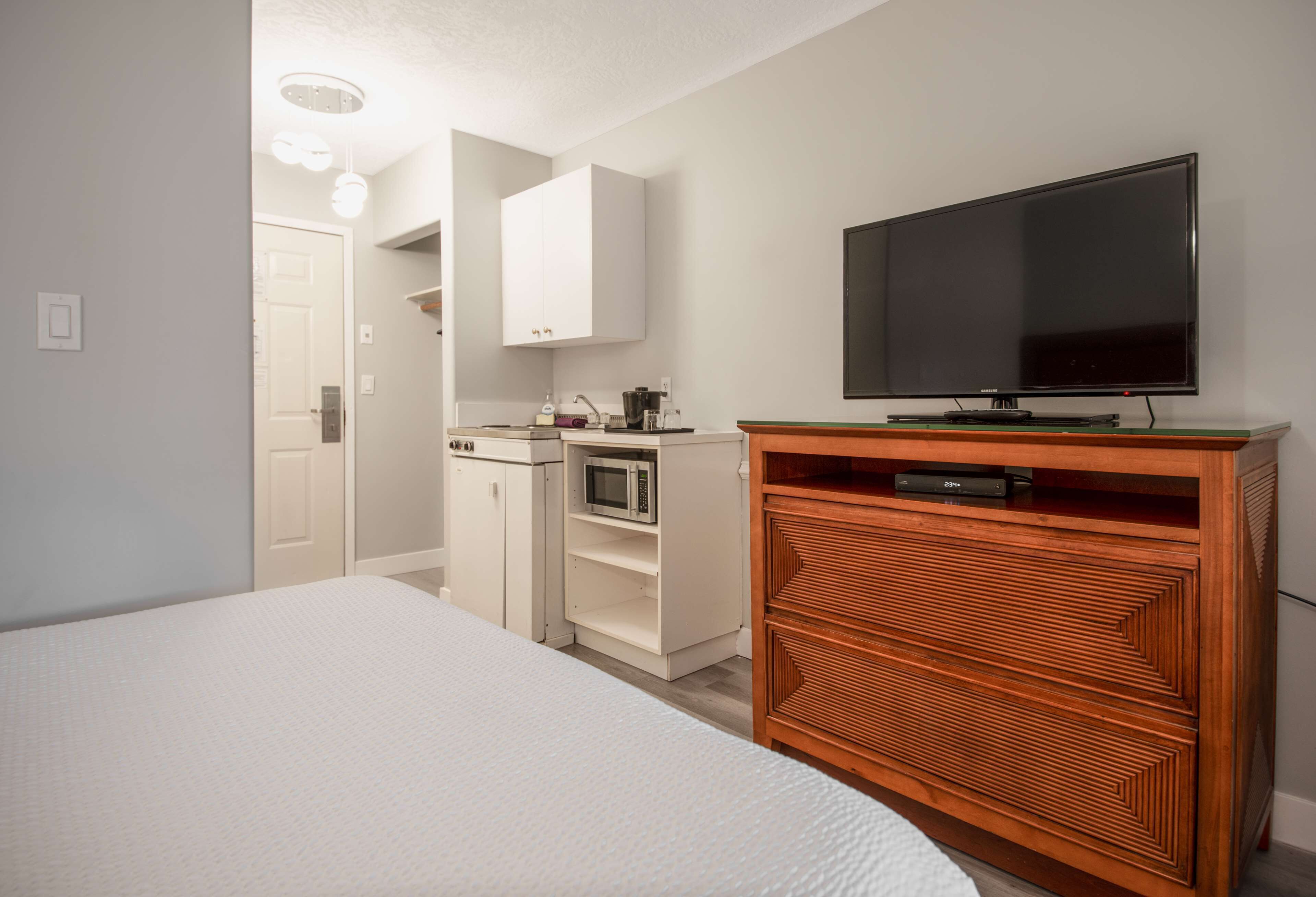 A bedroom with a bed, a wooden dresser, a television, a microwave, and a door leading to a bathroom.