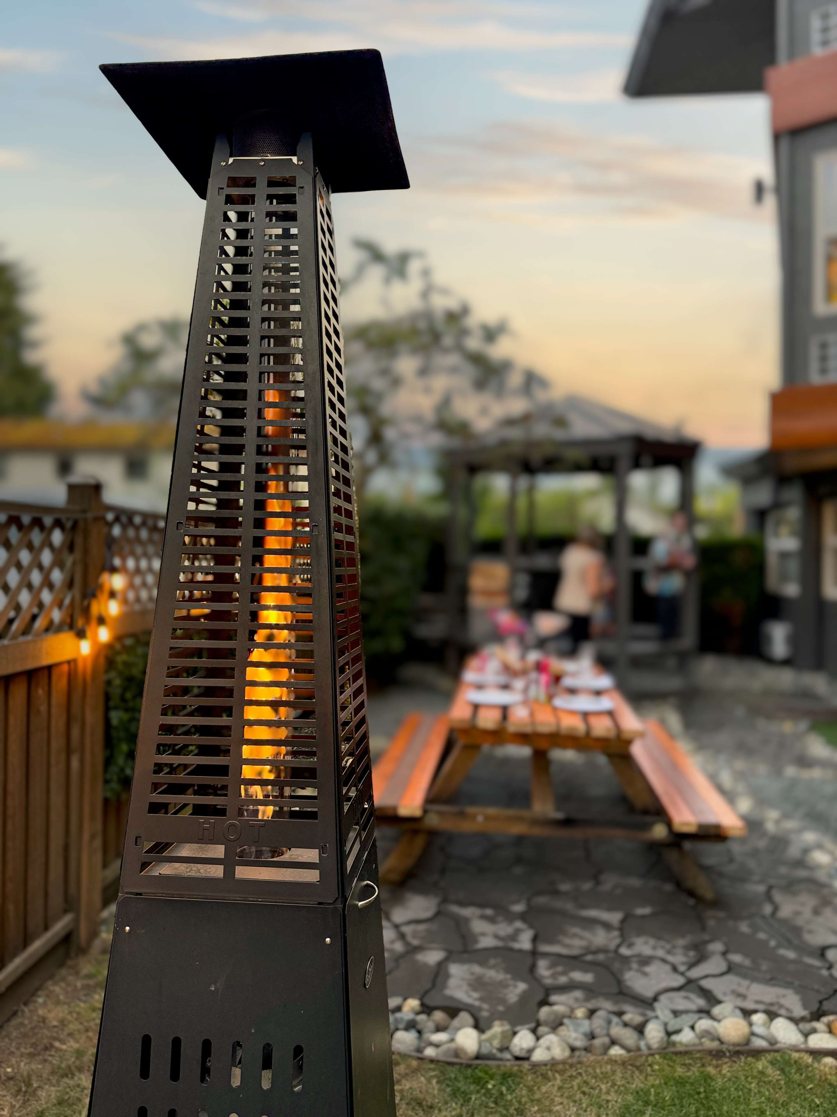 Un calentador metálico para exteriores con diseño de listones, una mesa de picnic y una valla de madera en un patio trasero.