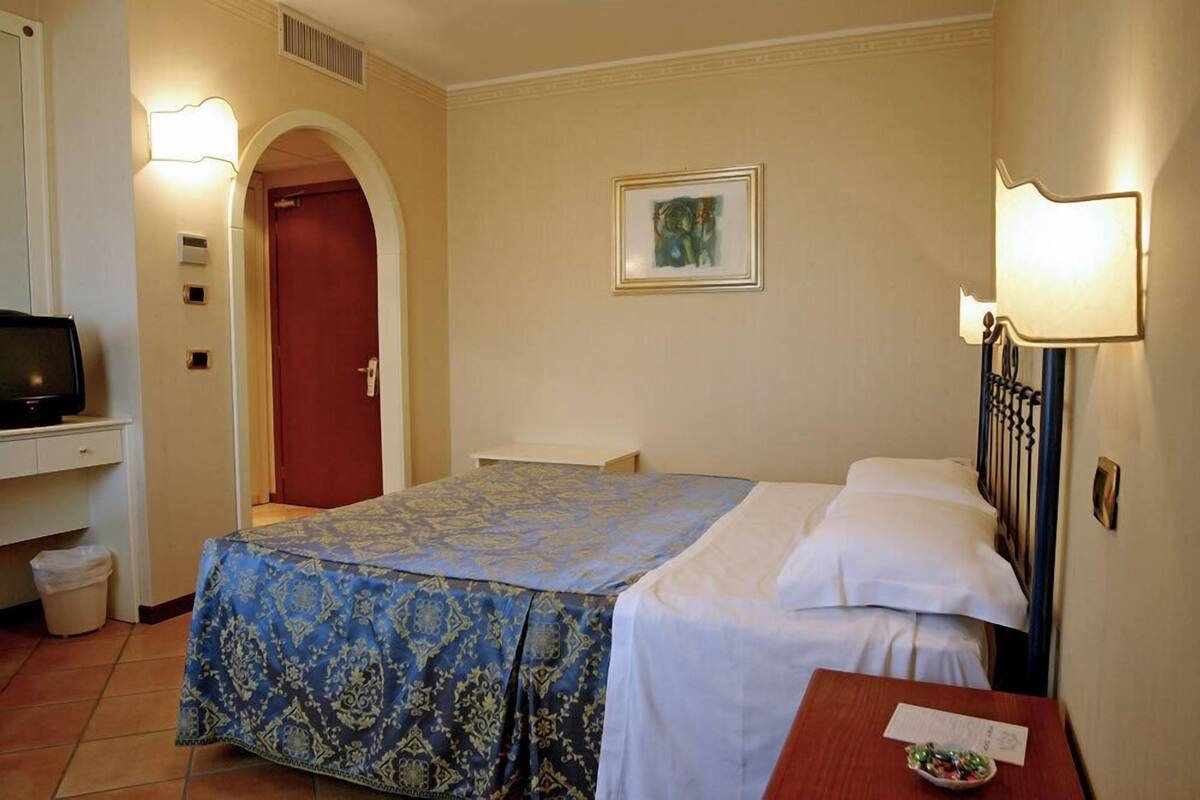 Habitación de hotel con cama, escritorio con televisor, puerta roja y lámpara de pared.