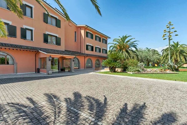 A multi-story building with a paved courtyard, palm trees, and a clear sky. - Hotel Villa Luigi (Martinsicuro)