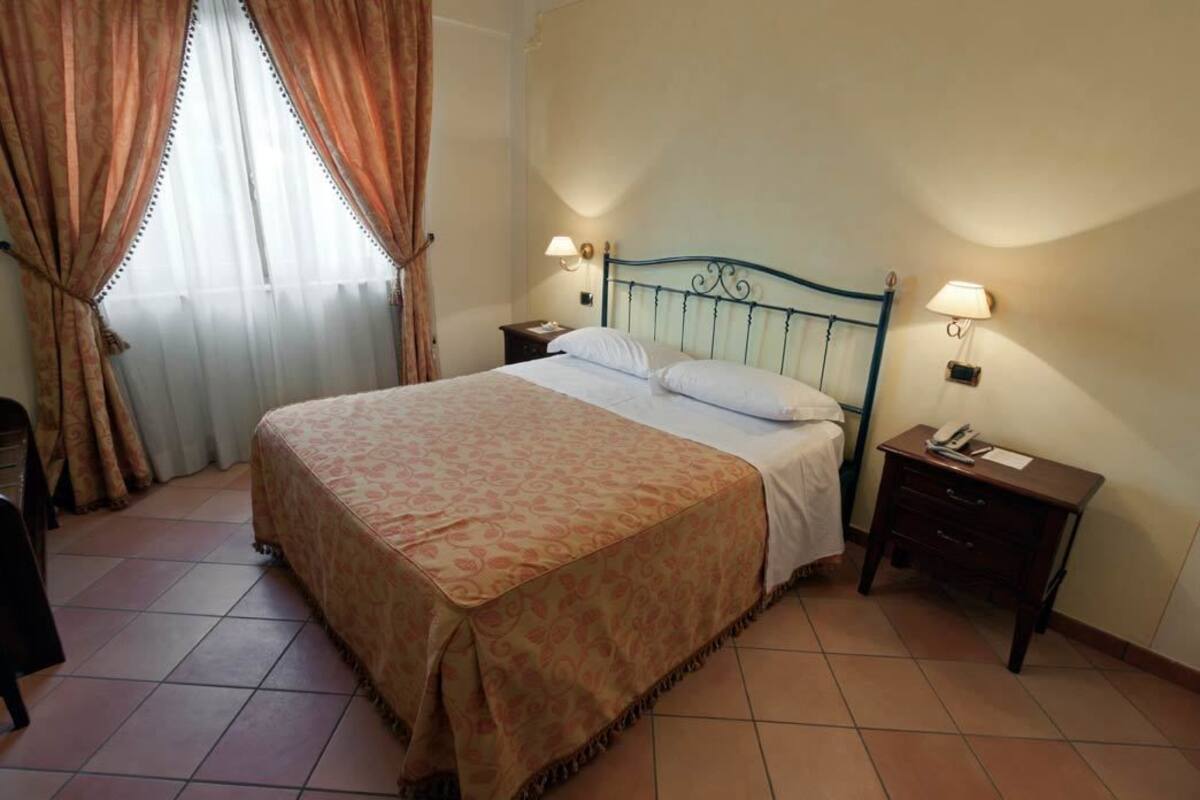 Una cama bien hecha con una colcha estampada, dos mesitas de noche con lámparas y una ventana con cortinas.