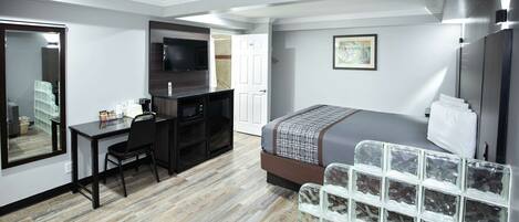 A compact hotel room with a bed, a desk, a chair, a TV, and a storage unit.