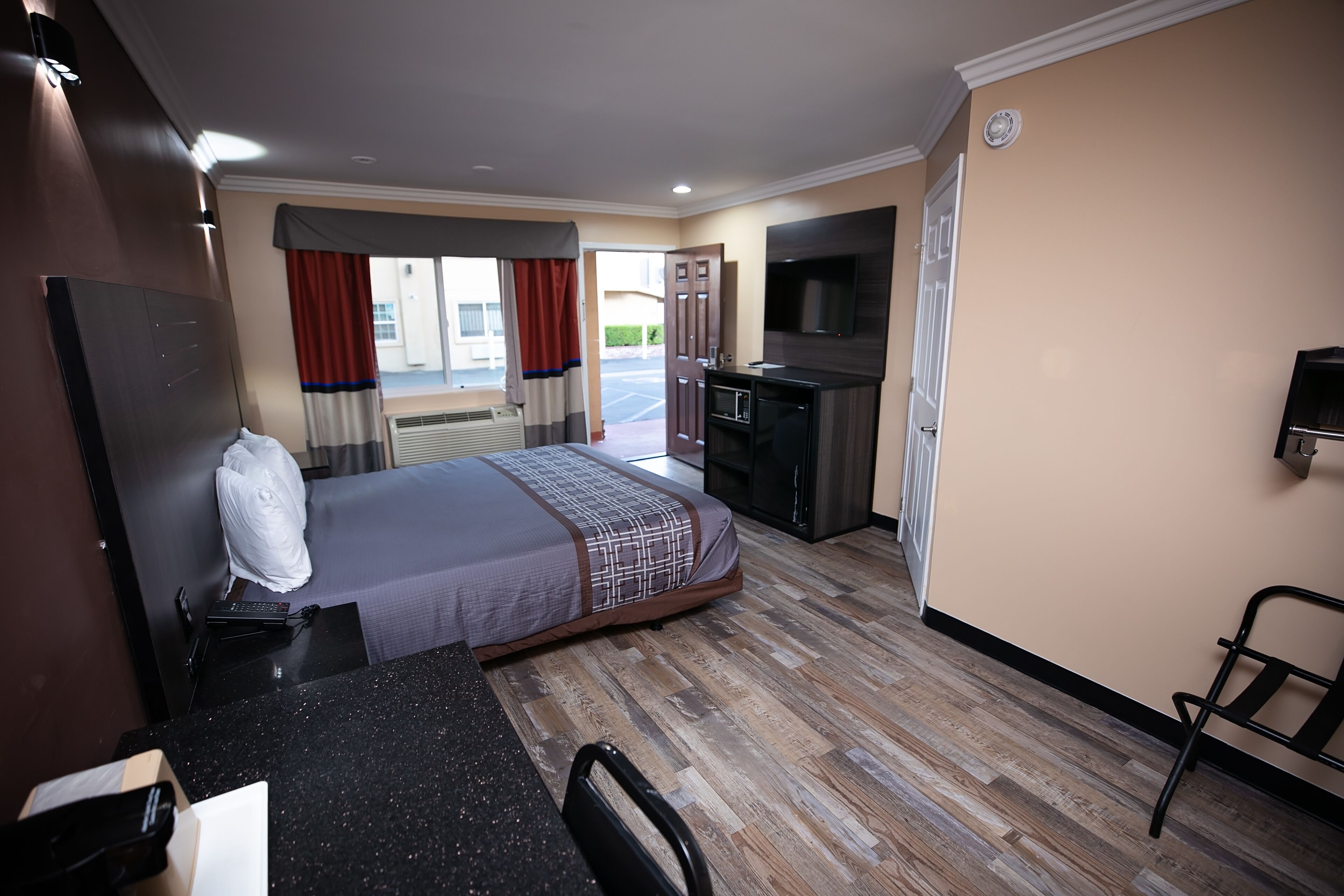 Habitación de hotel con cama, escritorio con silla, televisión, kitchenette y ventana con cortinas.