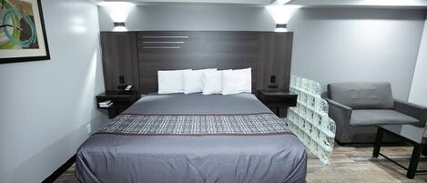 Une chambre d’hôtel moderne avec un grand lit, un fauteuil recouvert d’un tissu gris et un plancher en bois.