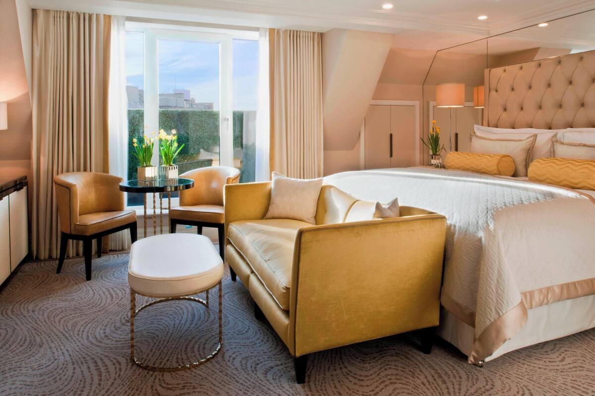 A hotel room with a large bed, two armchairs, a small table, and a view of the city through large windows.