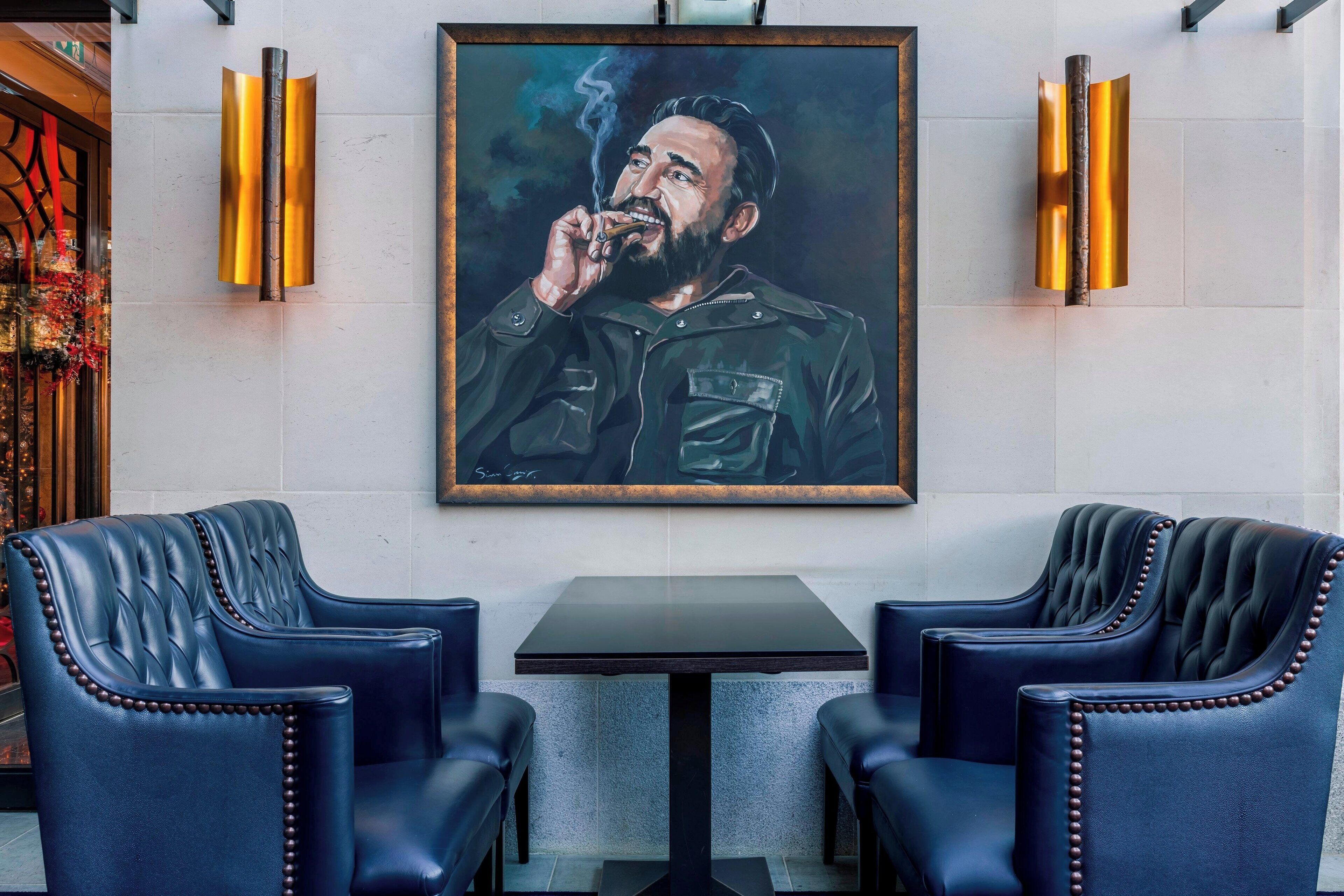 A framed portrait of a man in a leather jacket, two upholstered chairs, and a small table in a room with a tiled wall.