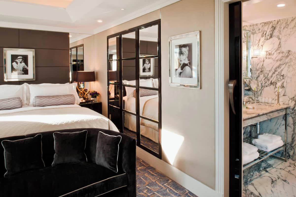A hotel room with a large bed, a dark sofa, a mirror, and a bathroom with a marble wall and a sink.