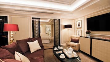 Une chambre d’hôtel avec un canapé rouge, un fauteuil beige, une table basse avec du thé et des pâtisseries, une télévision et un lit à l’arrière-plan.
