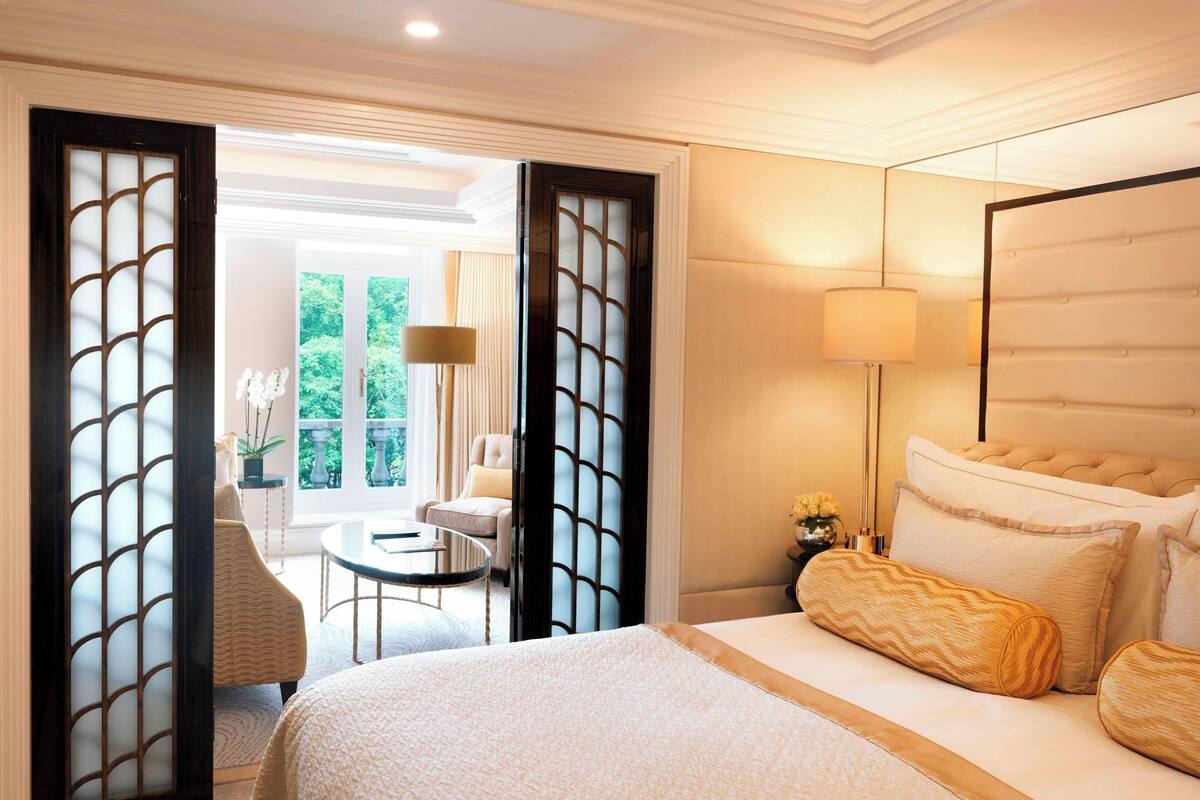 A hotel room with a bed, a bedside table, a lamp, and a view of the outdoors.