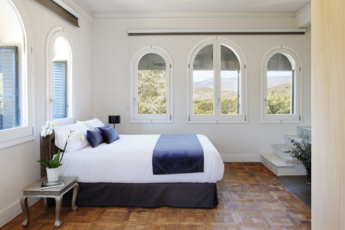 A bedroom with a large bed, three arched windows, a small table with a plant, and a staircase.