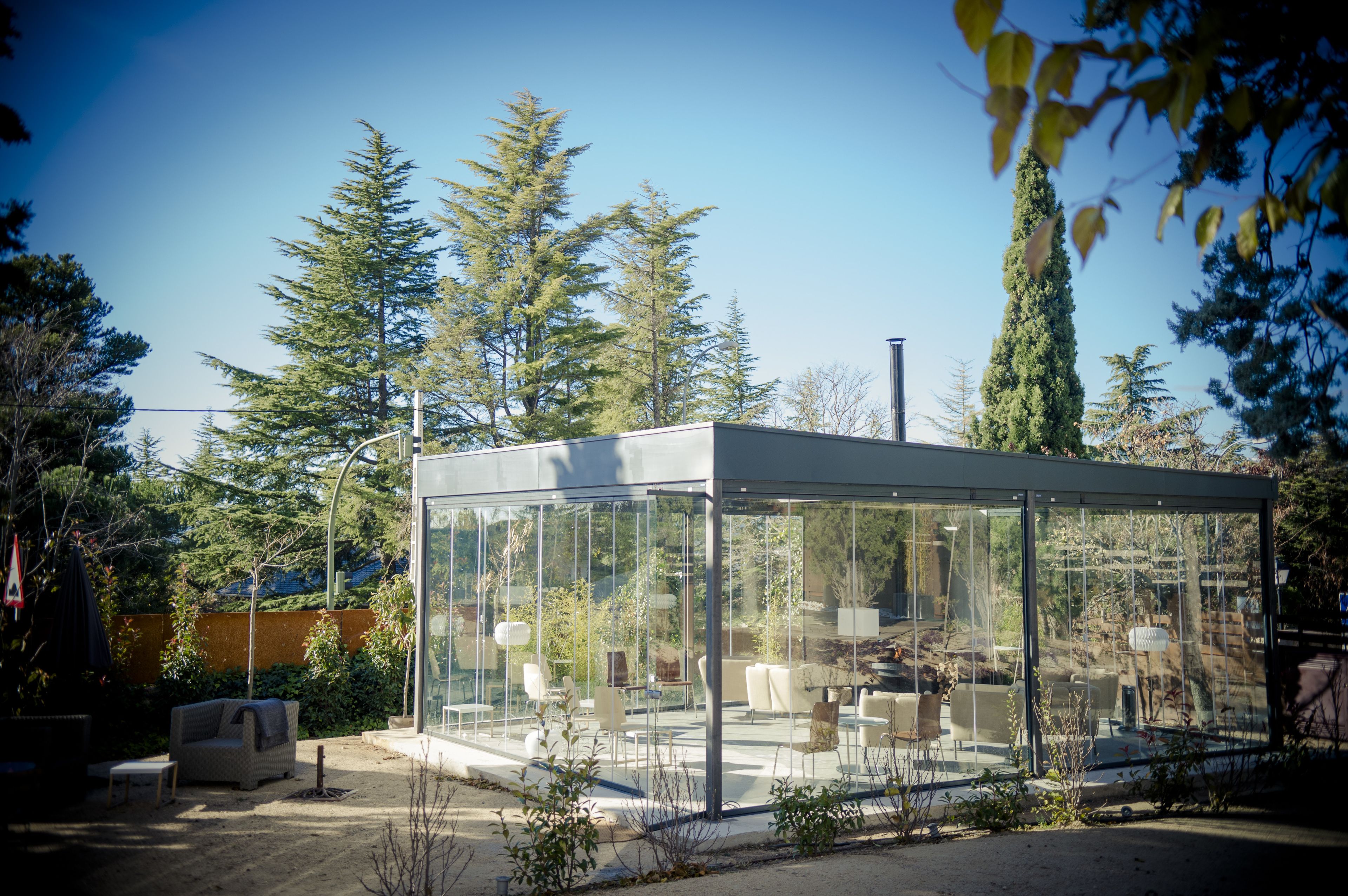 Un salón exterior moderno, con paredes de vidrio, con asientos y vistas a árboles circundantes.