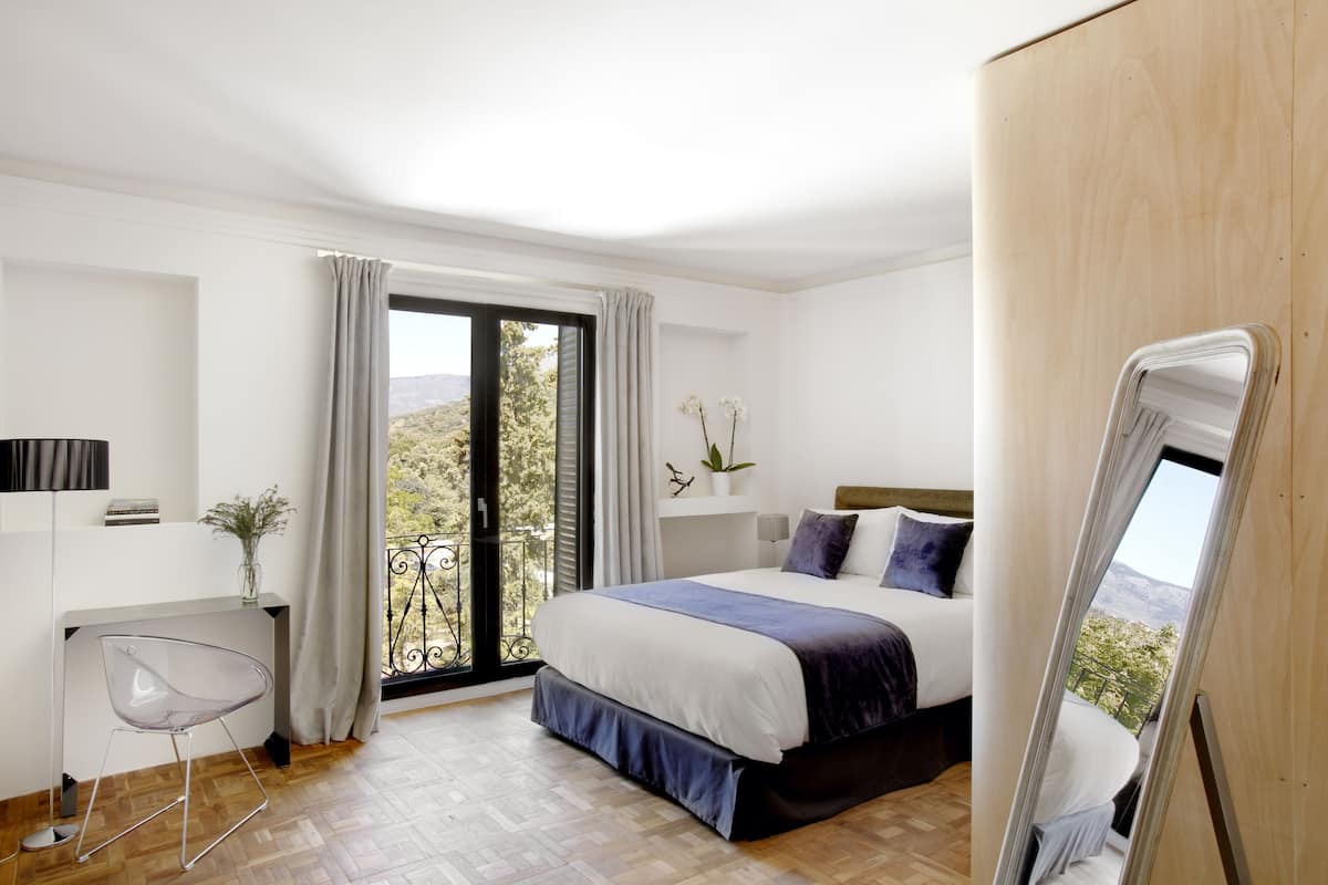 A modern bedroom with a large bed, a desk, and a balcony with a view.