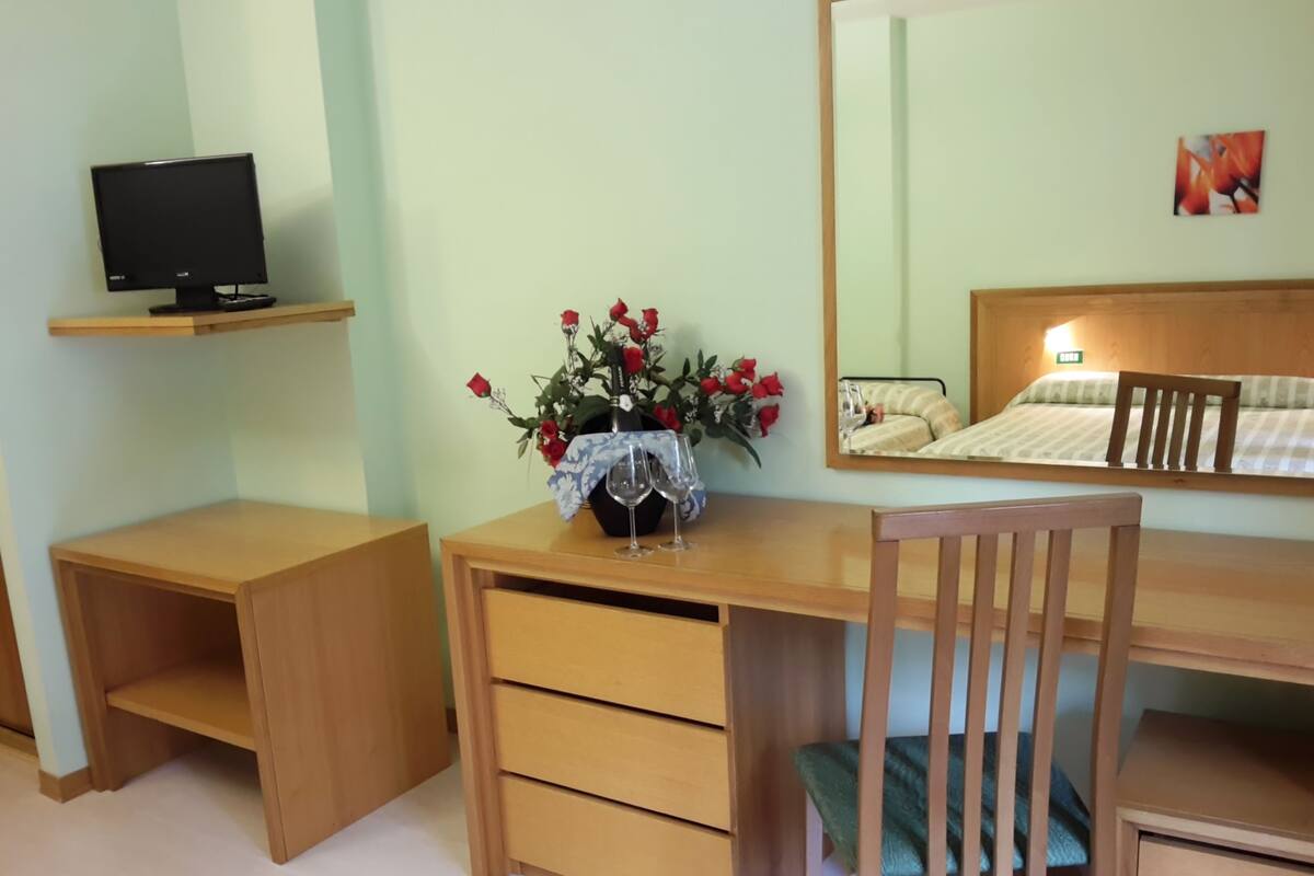 A room with a wooden desk, a chair, a TV on a shelf, and a bed with a headboard.