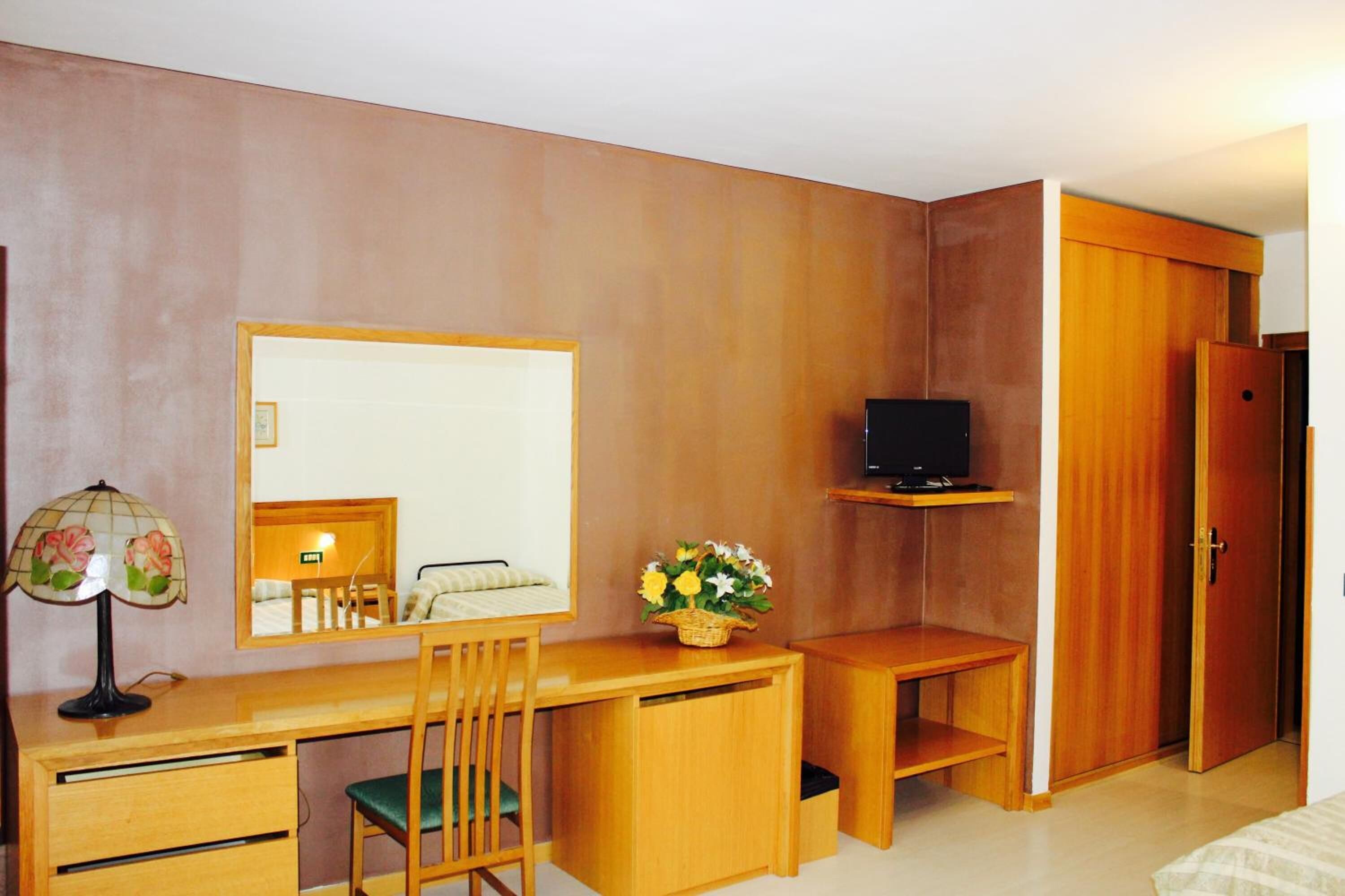 A compact room with a wooden desk, a chair, a television on a shelf, and a wardrobe.
