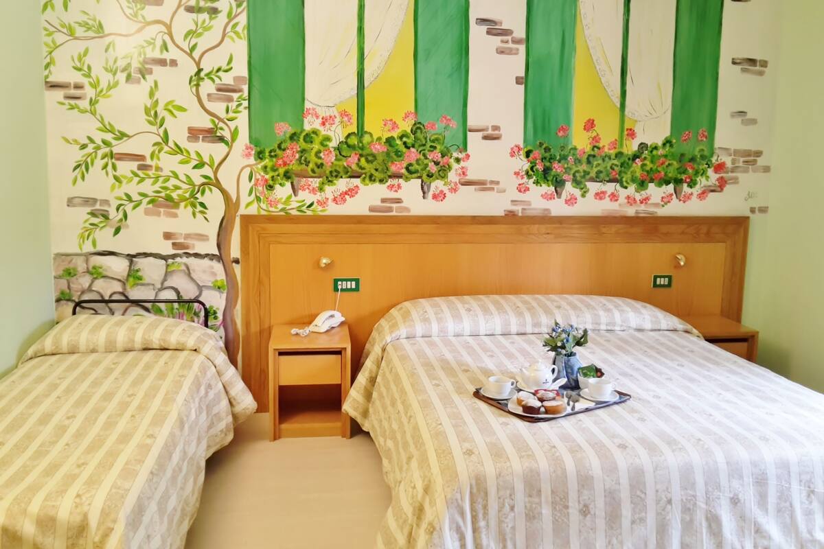A hotel room with two beds, a wooden headboard, a nightstand with a phone, and a wall mural with a tree and flowers.