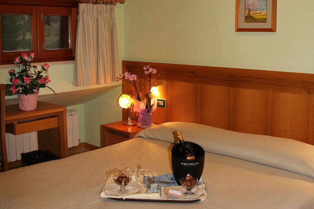 A bedroom with a wooden ceiling, a bed with a tray of breakfast items, a nightstand with a lamp, and a window with curtains.