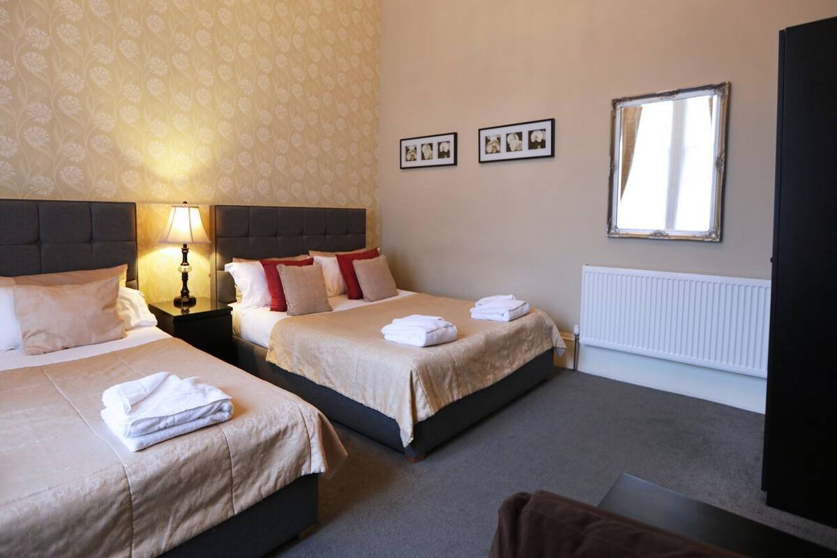 A hotel room with two beds, a nightstand with a lamp, a radiator, and framed pictures on the wall.