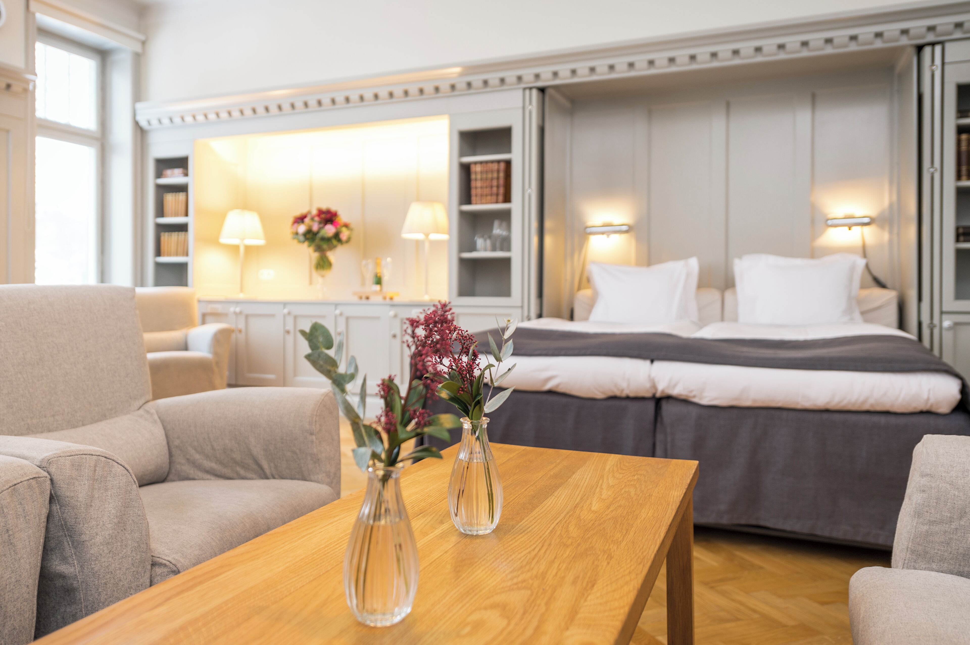 A modern hotel room with a wooden coffee table, two vases with flowers, a bed with white and gray bedding, and a seating area with a sofa and armchair.
