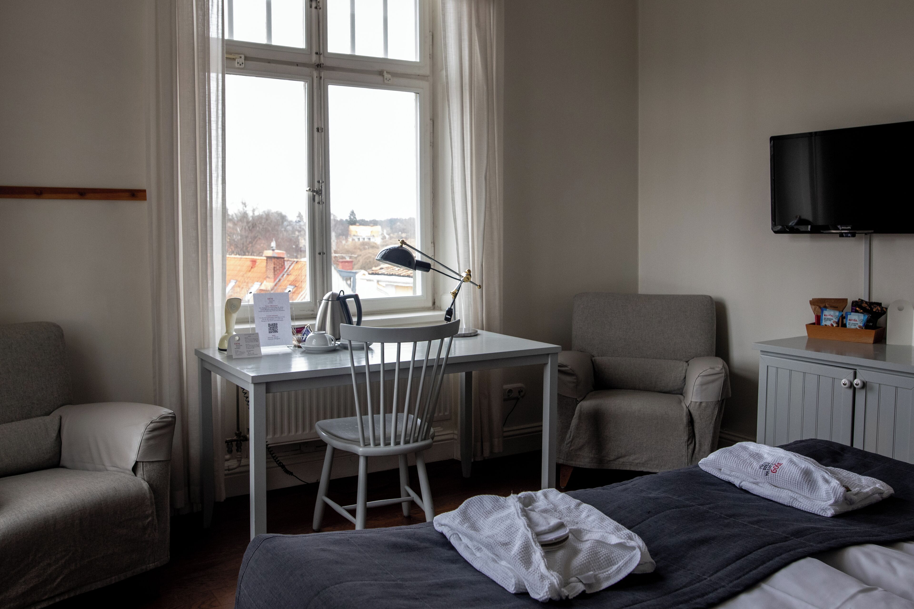 Une chambre d’hôtel équipée d’un bureau, de deux chaises, d’un lit, d’une télévision et d’une fenêtre donnant sur des immeubles.