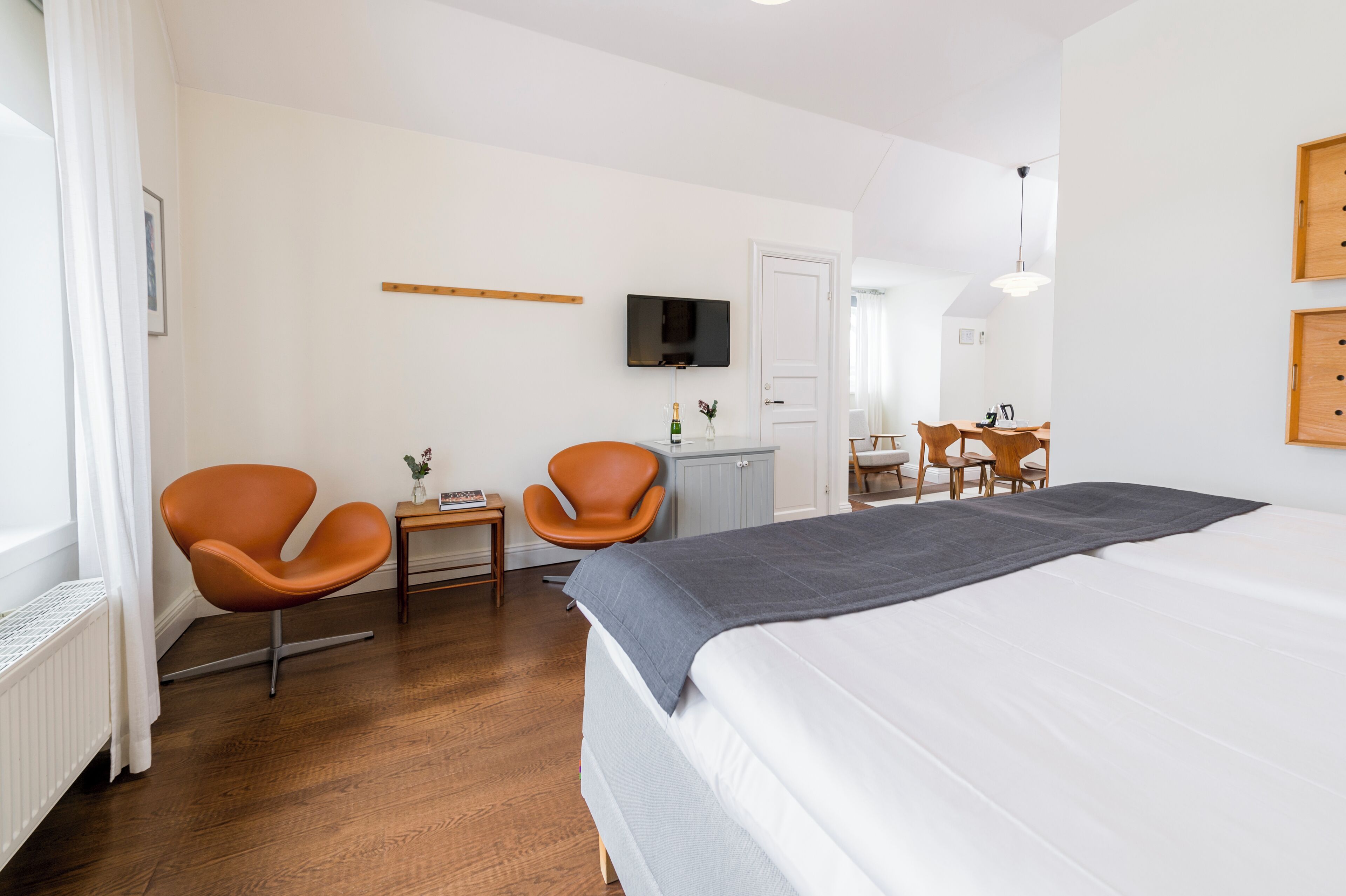 Een moderne hotelkamer met een groot bed, twee oranje stoelen, een klein tafeltje met een fles, een televisie aan de muur en een houten deur.