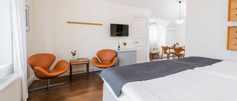 Een moderne hotelkamer met een groot bed, twee oranje stoelen, een klein tafeltje met een fles, een televisie aan de muur en een houten deur.
