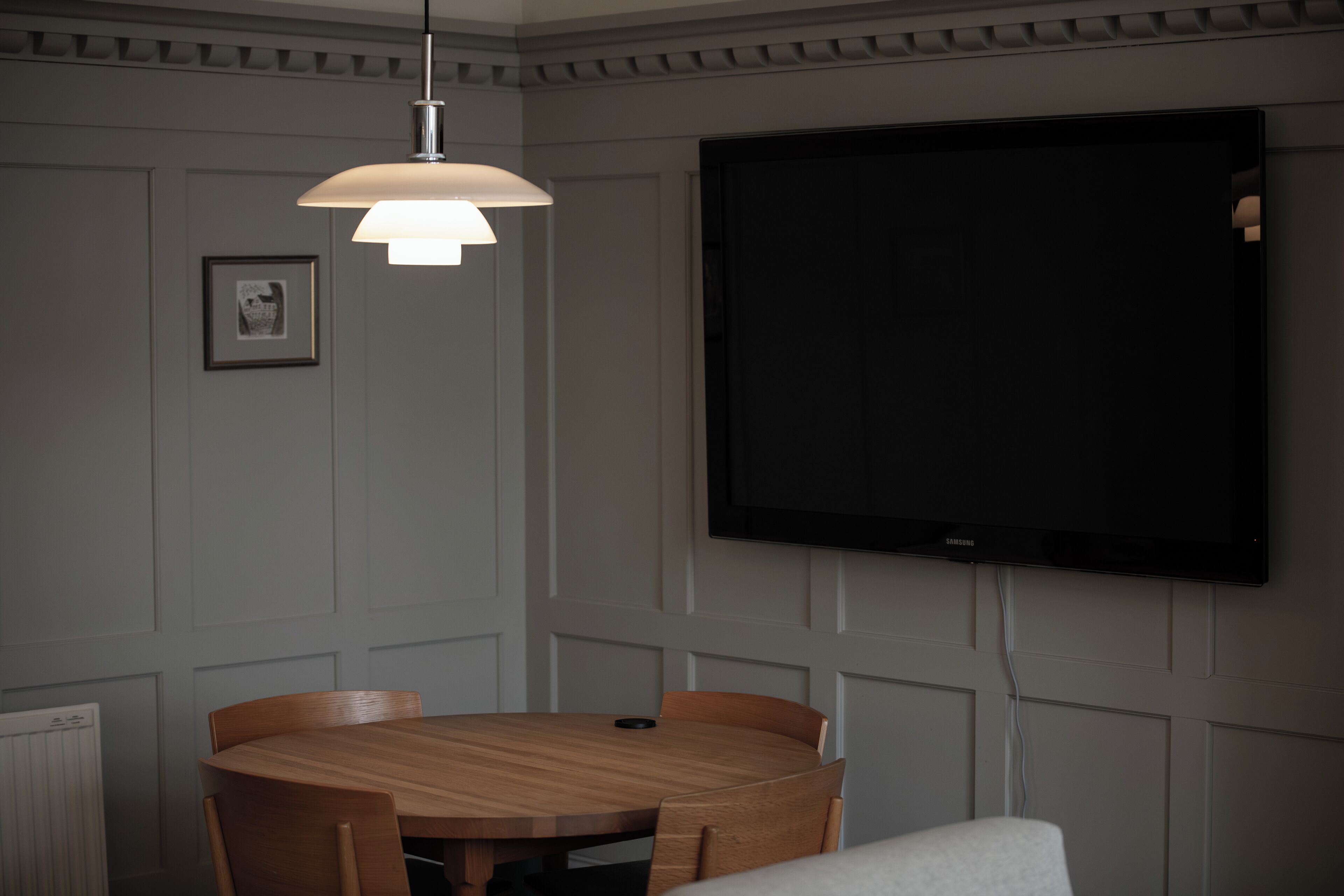 A dining area with a round wooden table, chairs, a pendant light, and a flat-screen TV mounted on the wall.