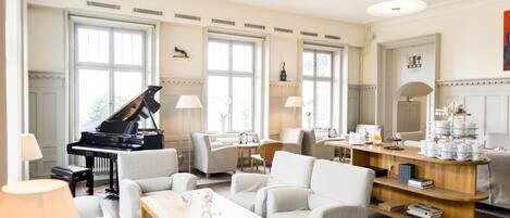 Ein geräumiges Wohnzimmer mit einem Flügel, bequemen Sitzgelegenheiten, einer Kaffeestation und großen Fenstern.