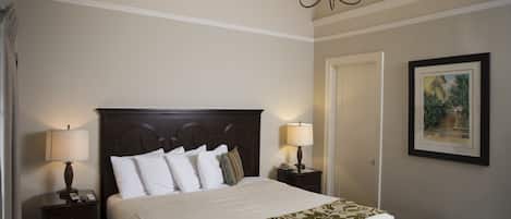 Un lit bien fait, avec du linge de lit blanc et un couvre-lit à motifs floraux, deux lampes de chevet, une tête de lit en bois foncé et un tableau encadré au mur.
