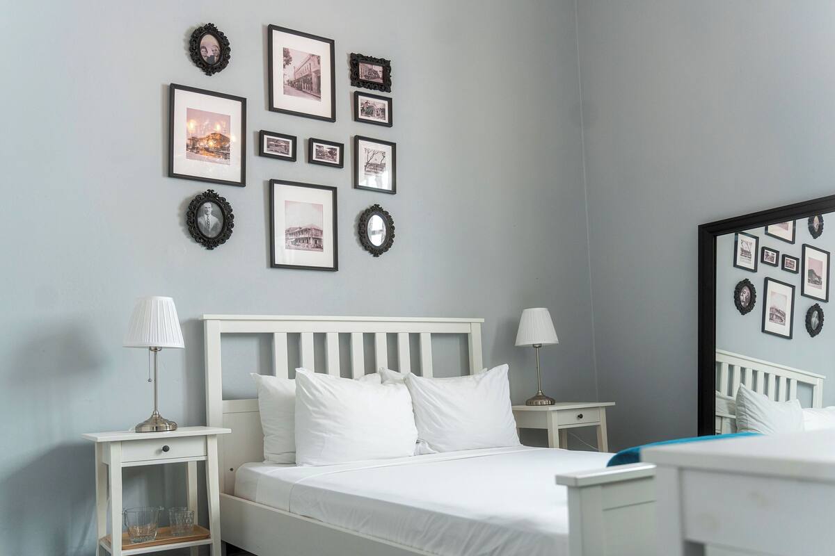 A neatly arranged bedroom with a bed, two bedside tables, a nightstand, and a wall adorned with framed pictures.