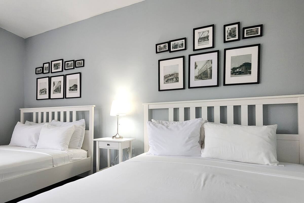 A hotel room with two beds, a bedside table with a lamp, and a wall with framed pictures.