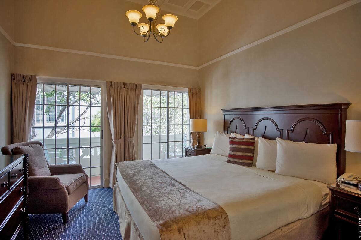 A hotel room with a large bed, a dresser, a chair, a nightstand with a lamp, and a chandelier.
