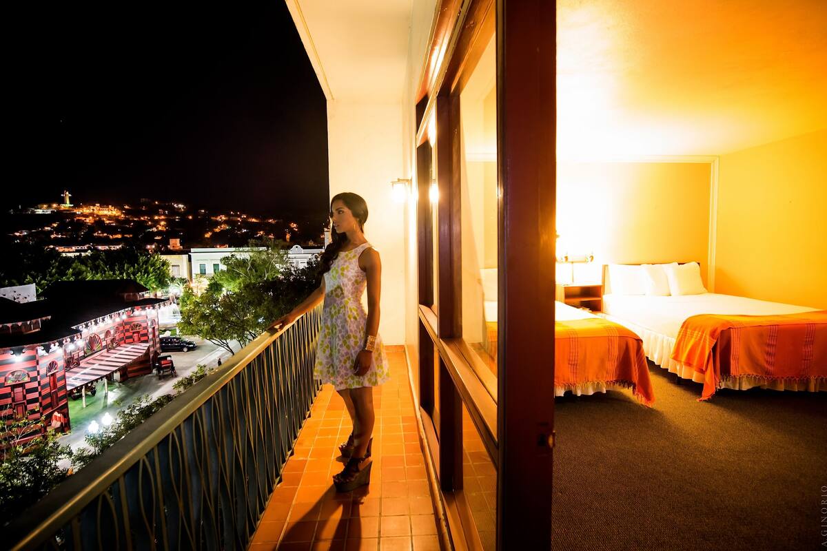 A balcony with a view of a cityscape at night, a bed with a white comforter, and a nightstand with a lamp.