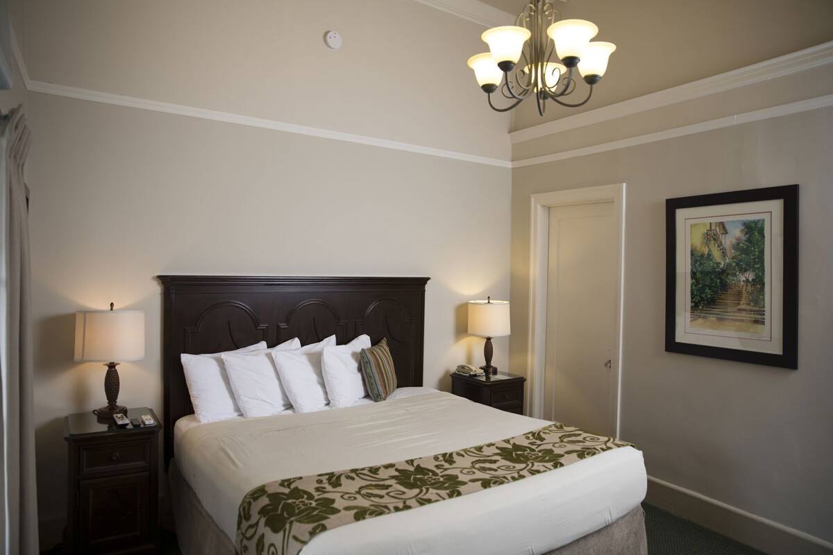 A hotel room with a large bed, two bedside tables with lamps, a chandelier, a framed picture on the wall, and a door.