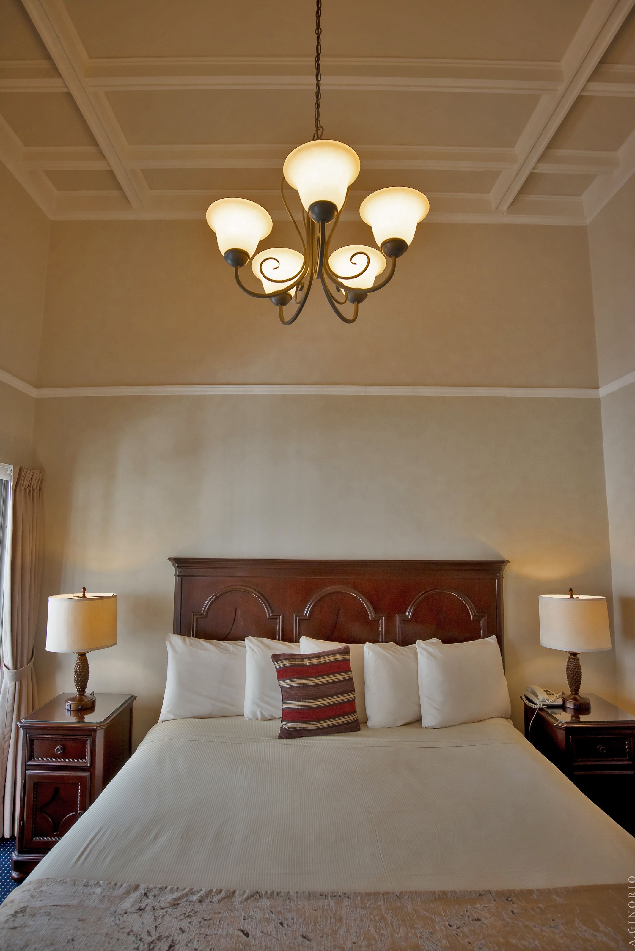 A hotel room with a large bed, a wooden headboard, two bedside tables with lamps, and a chandelier.