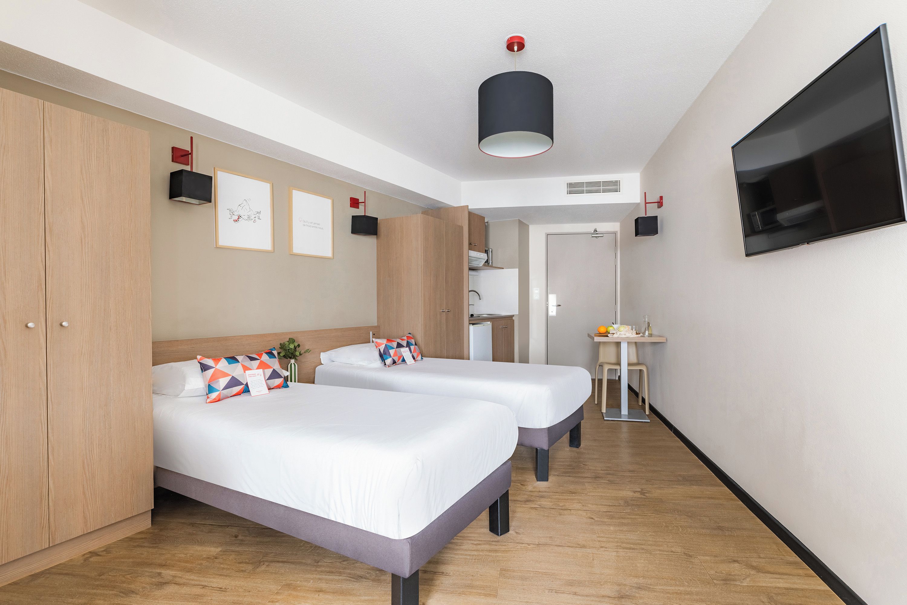 Ein Hotelzimmer mit zwei Betten, einem Fernseher, einem kleinen Tisch und einer Küchenzeile.
