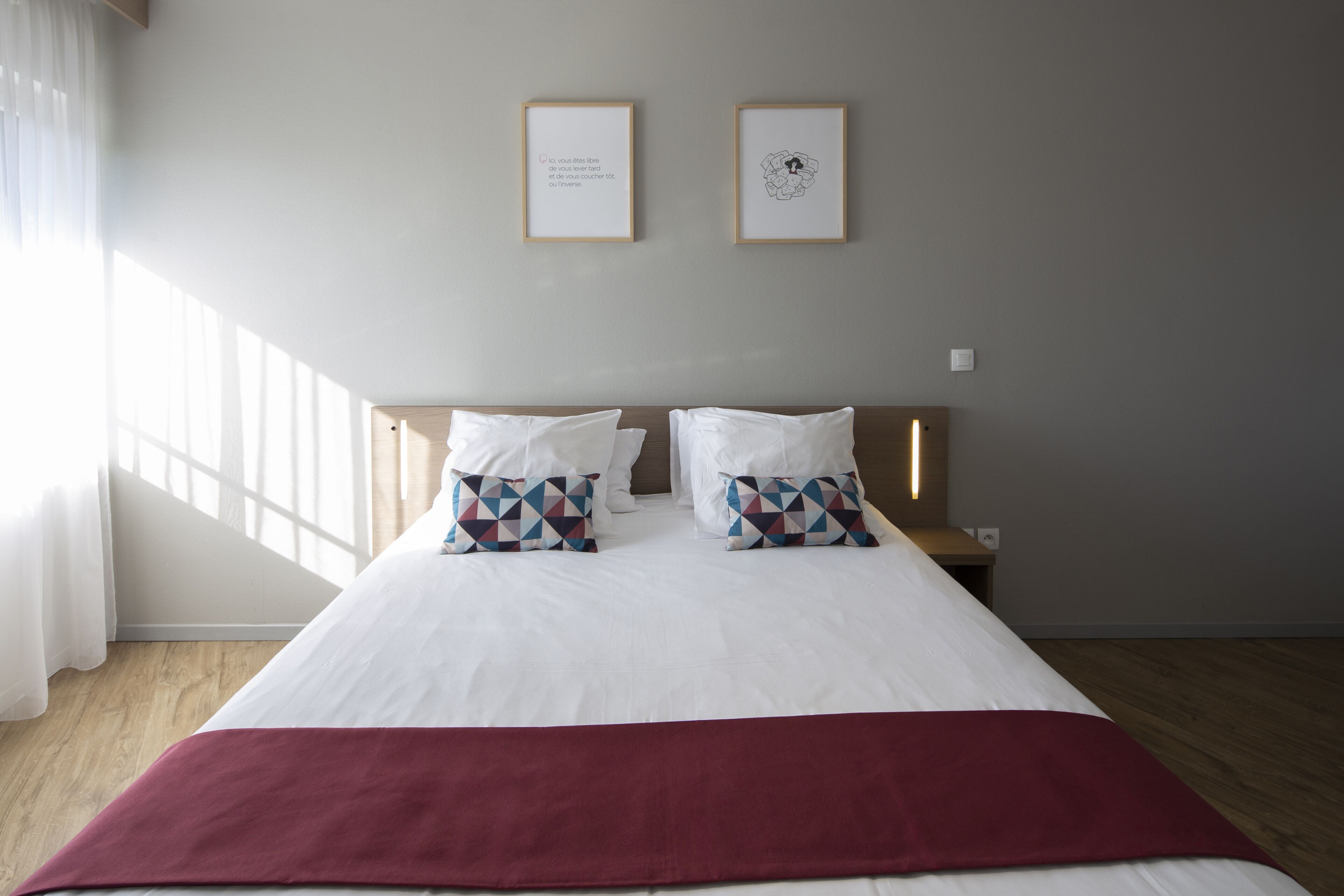 Una cama bien hecha con ropa blanca y dos almohadas estampadas, un cabecero de madera y dos cuadros en la pared.