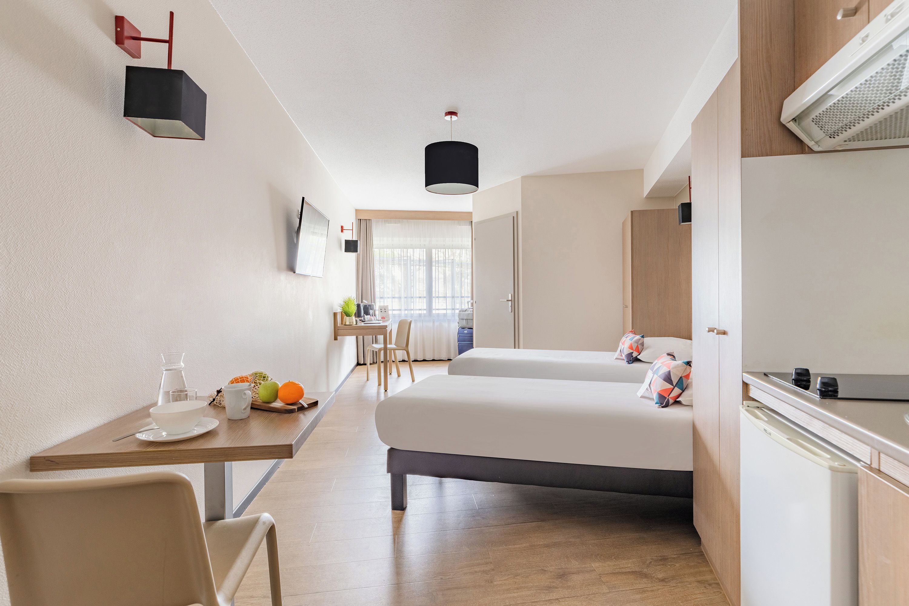 A hotel room with a bed, a desk, a chair, a small table with a cup and fruits, and a TV on the wall.