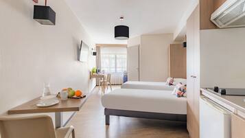 A hotel room with a bed, a desk, a chair, a small table with a cup and fruits, and a TV on the wall.