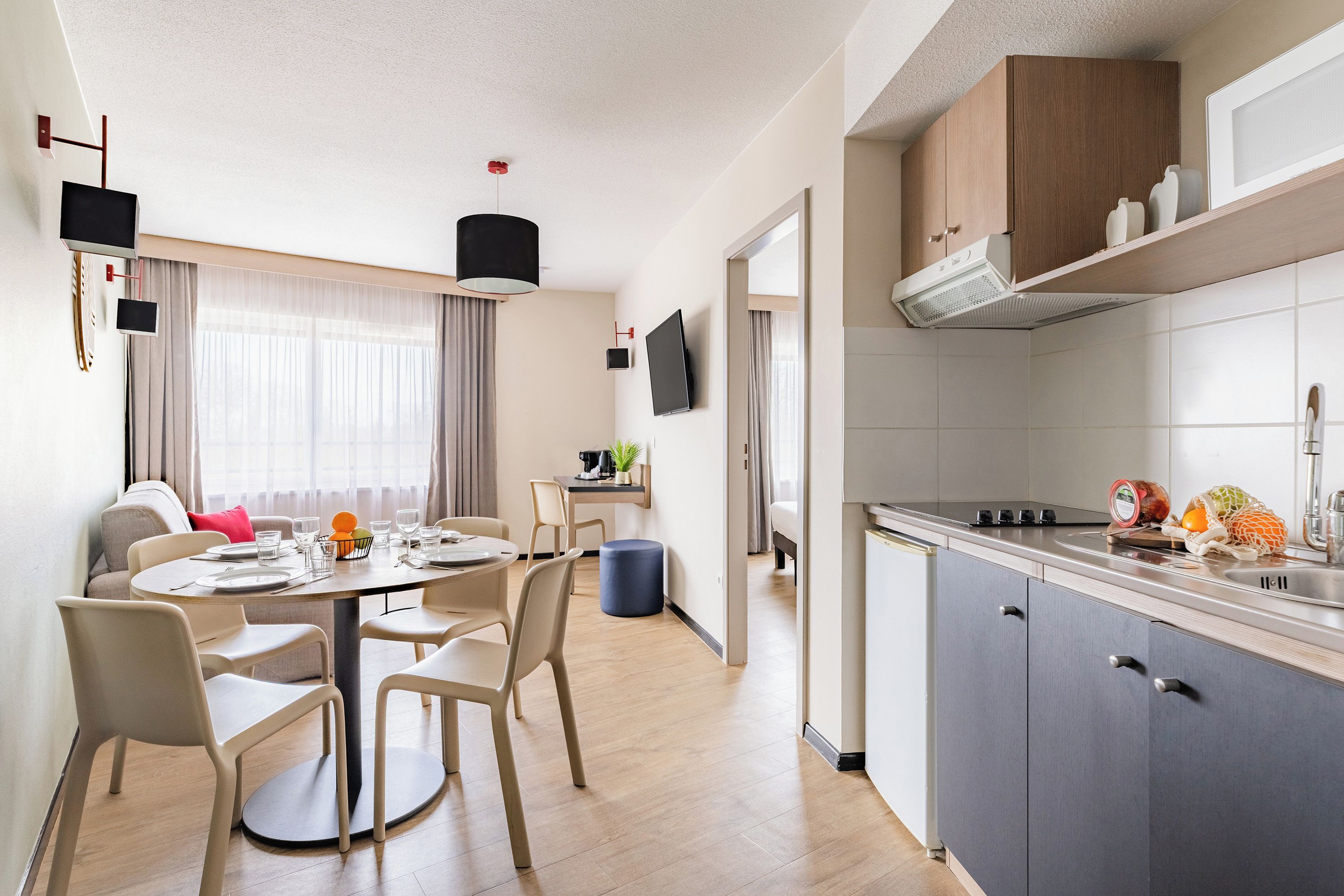 A modern kitchen with a built-in stove, a sink, and cabinets. There is a dining area with a round table and chairs, and a window with curtains.