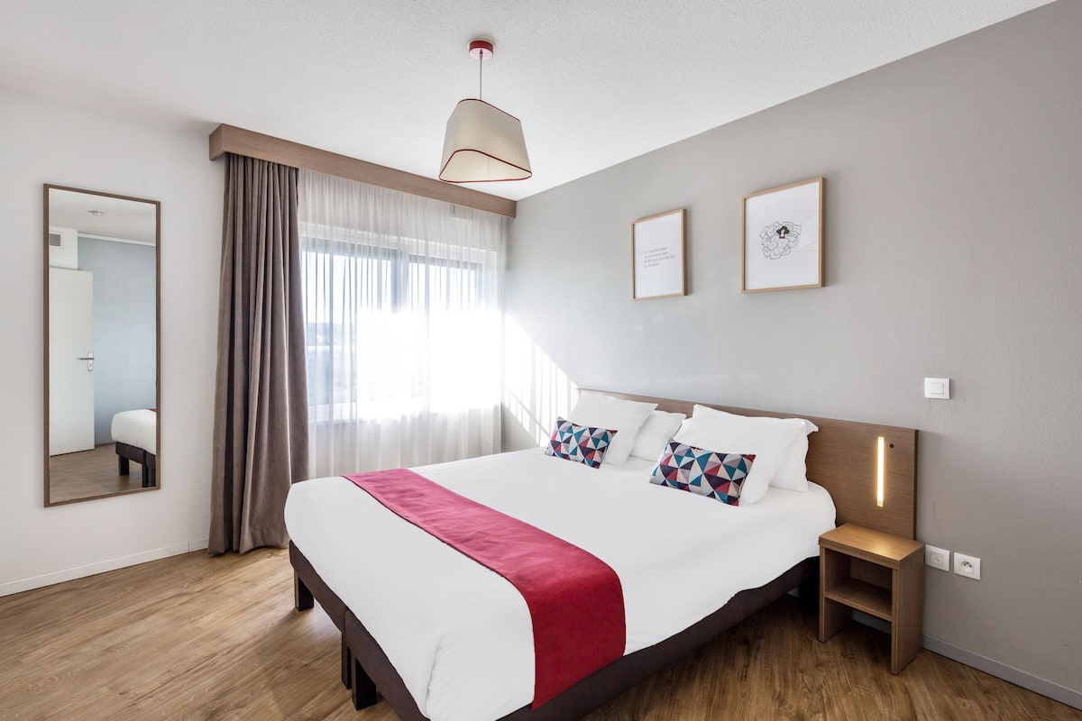 A modern hotel room with a large bed, a bedside table, a mirror, and a window with curtains.