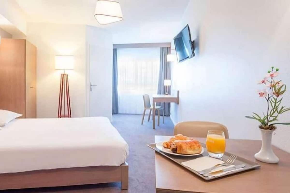 A hotel room with a bed, bedside table with breakfast, a TV, and a desk with a chair.