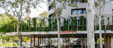 A modern hotel with a glass facade, outdoor seating, and a tree-lined entrance.