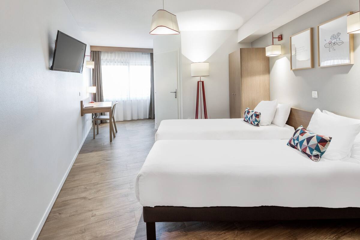 A hotel room with two beds, a desk, a chair, a TV, and a floor lamp.