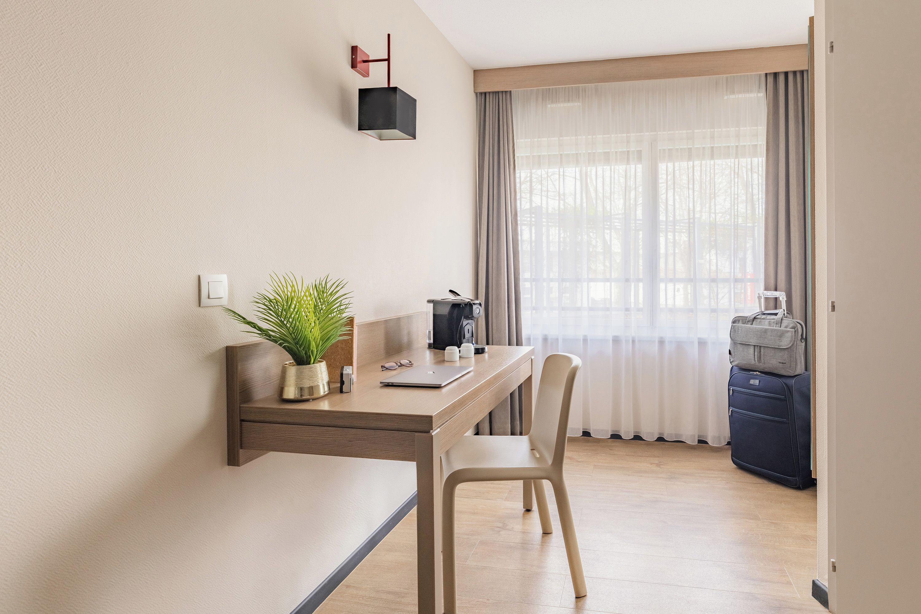 A small, modern room with a wooden desk, a chair, a potted plant, a coffee machine, and a suitcase.