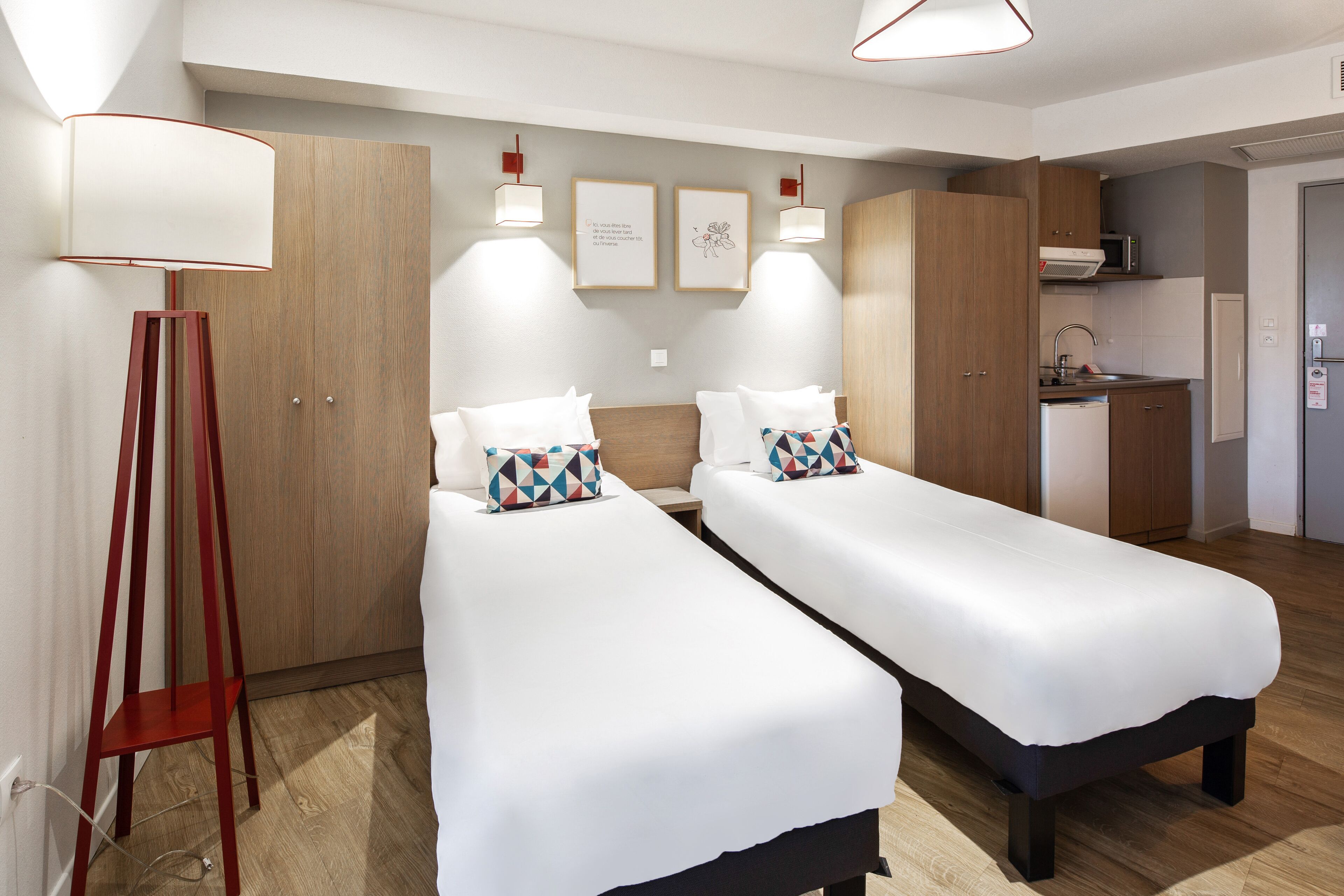 Habitación de hotel moderna con dos camas individuales, un armario de madera, una kitchenette y una lámpara de pie.