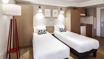 Habitación de hotel moderna con dos camas individuales, un armario de madera, una kitchenette y una lámpara de pie.