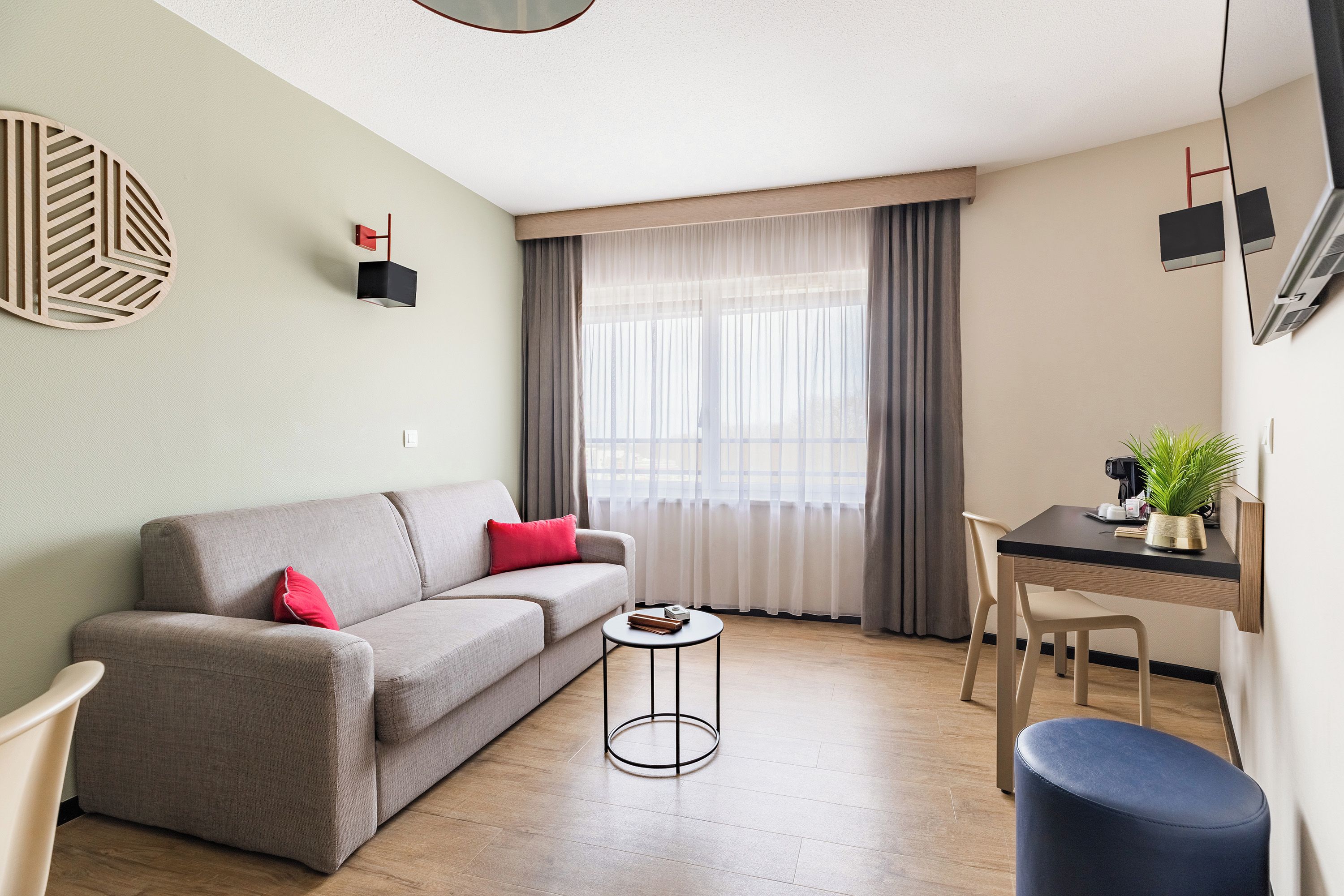 A modern living room with a sofa, a small round table, a dining area with a chair, and a large window with curtains.